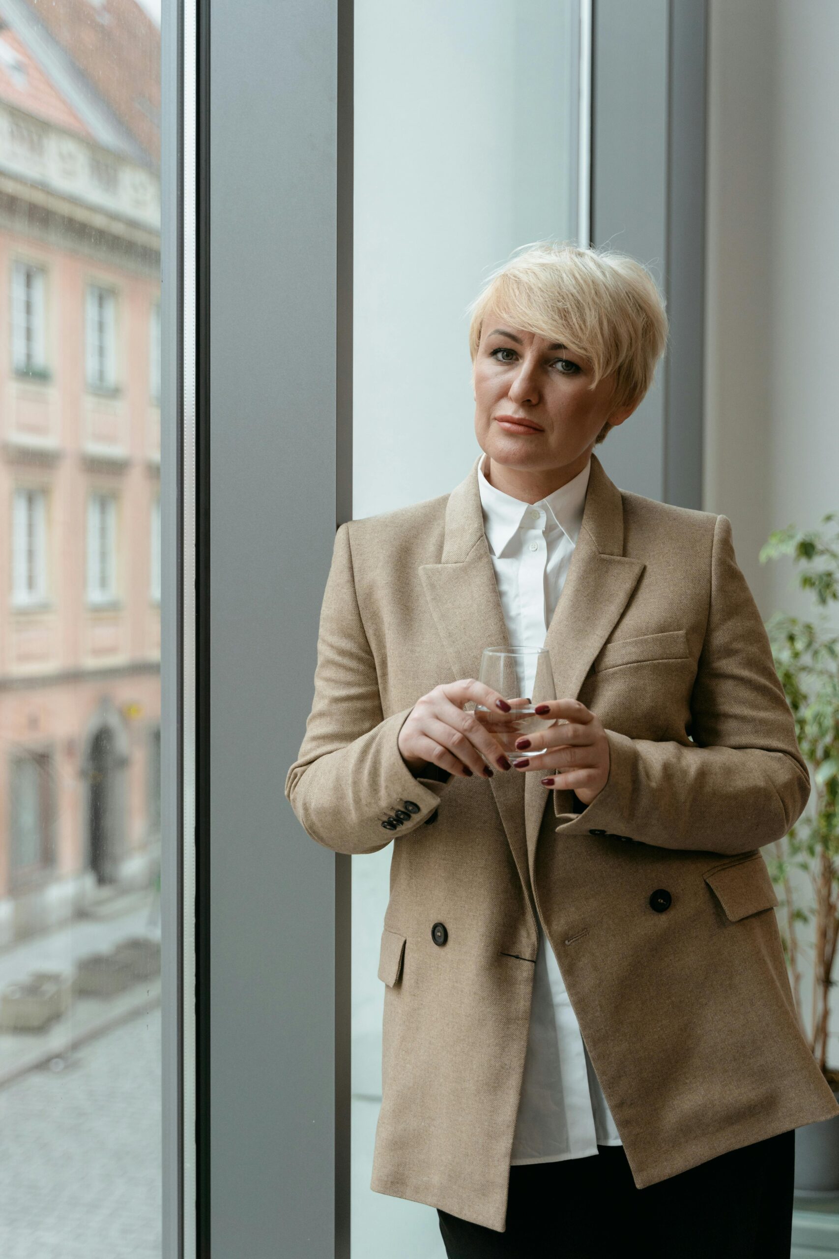 Caucasian businesswoman in formal attire standing by a window holding a glass.